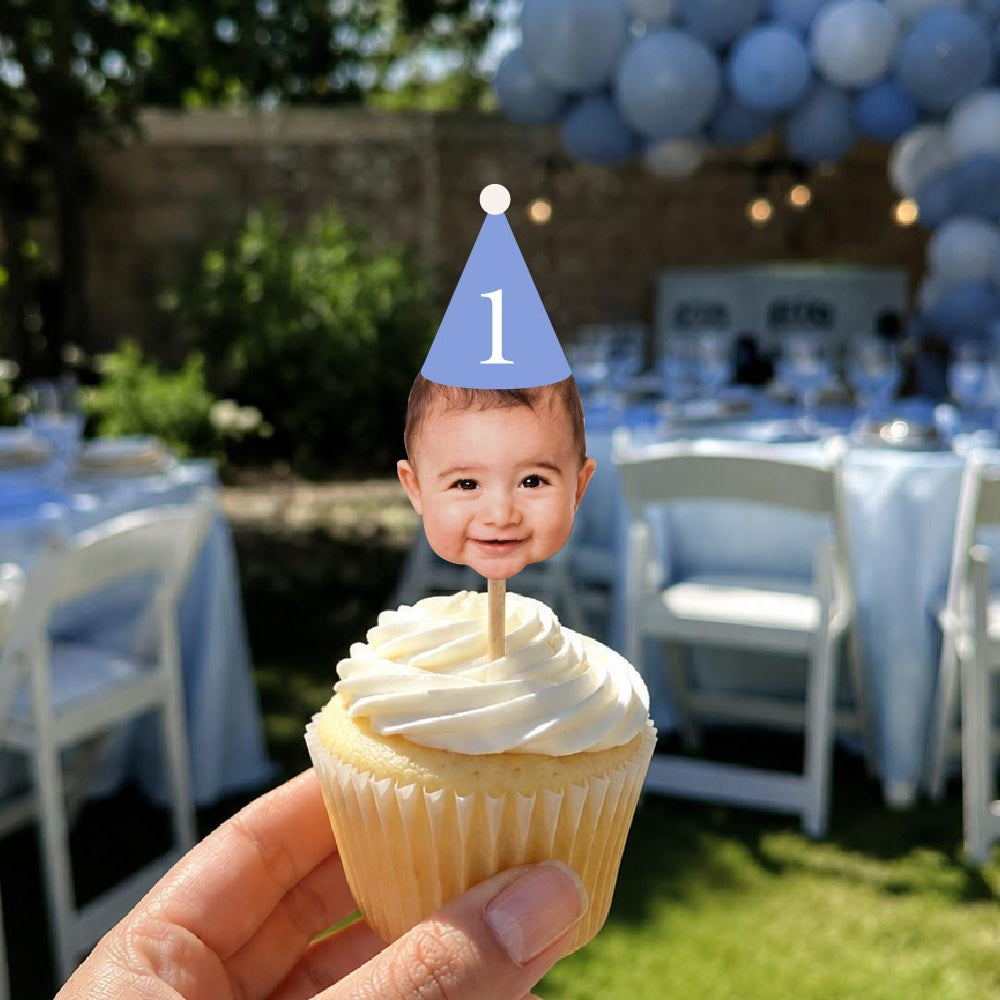 A hand holds a frosted cupcake with a custom photo topper showing a baby's face wearing a blue party hat with the number 1, set against a blurred outdoor birthday party background with tables and balloons.shop_this_look_amDhCa